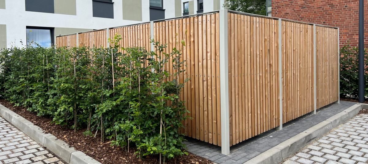 Sichtschutzzaun aus Holzlatten umgibt ein landschaftlich gestaltetes Eckgrundstück neben einem gepflasterten Gehweg.