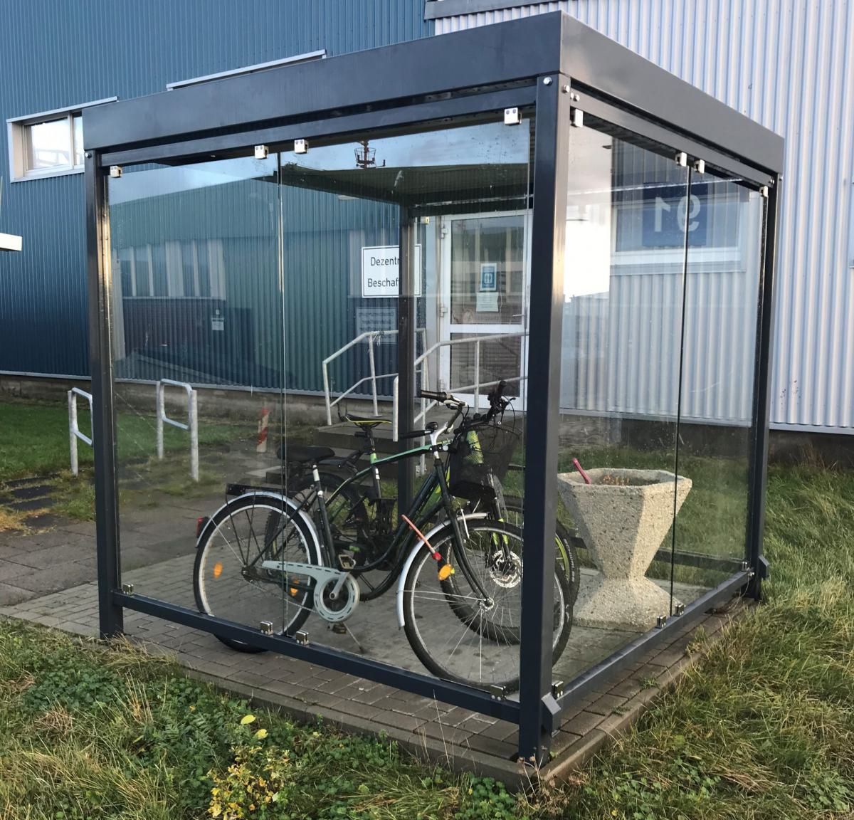 Das Fahrrad ist in einem gläsernen Fahrradunterstand neben einem blauen Gebäude geparkt.