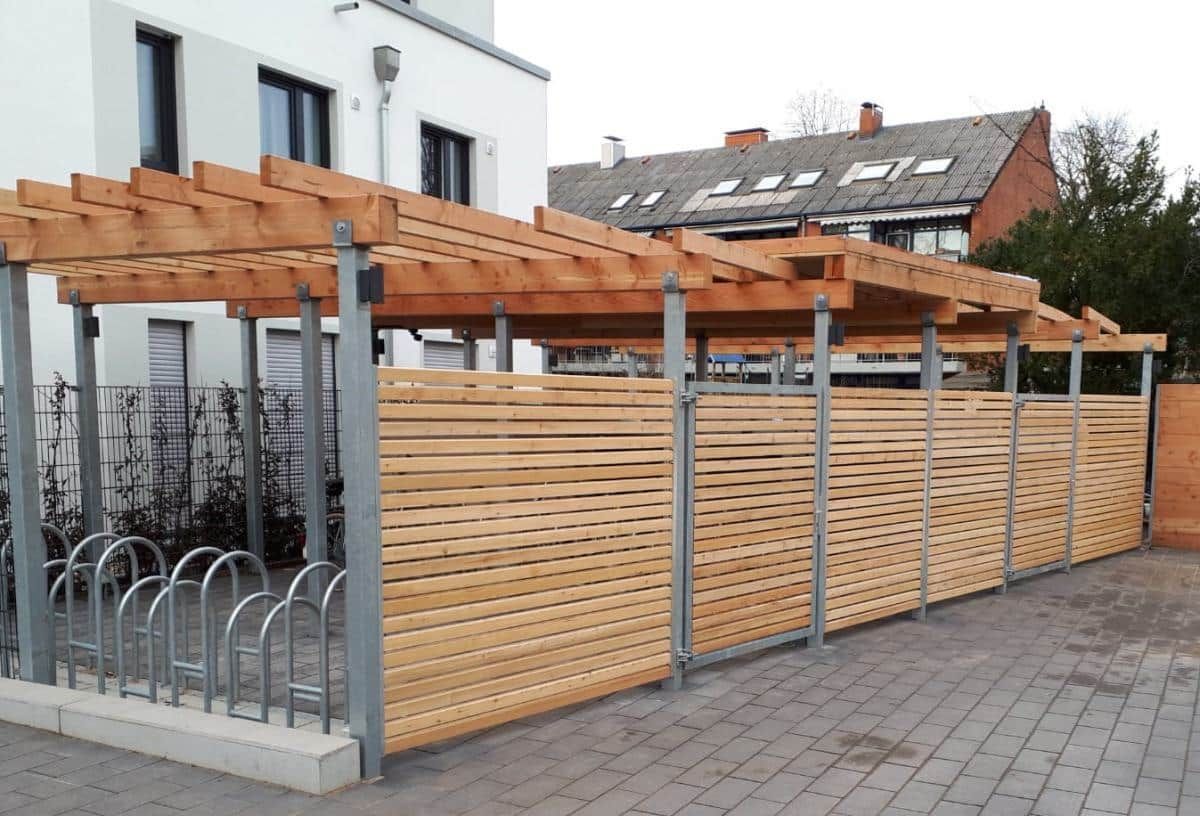 Eine hölzerne Pergola über einer umzäunten Terrasse neben einem weißen Gebäude mit grauen Pflastersteinen.