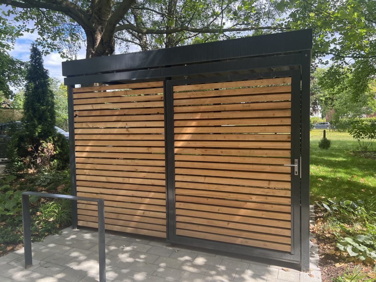 Holztor mit dunklem Metallrahmen und Lamellenpaneelen in einem grünen Parkbereich im Freien