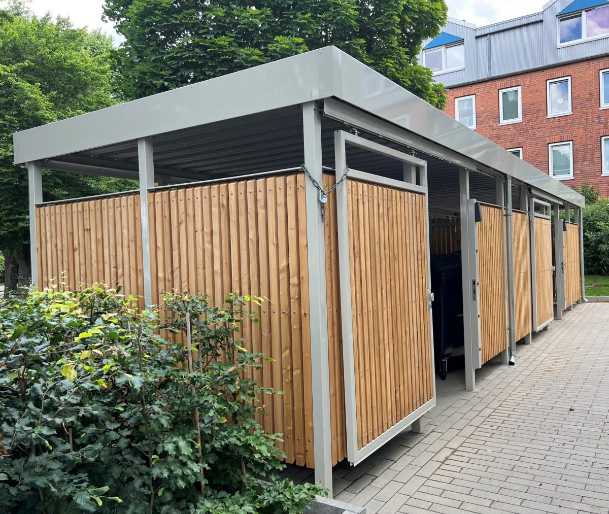 Hölzerner Fahrradunterstand mit grauem Dach neben einem Backsteingebäude und Bäumen entlang eines gepflasterten Weges