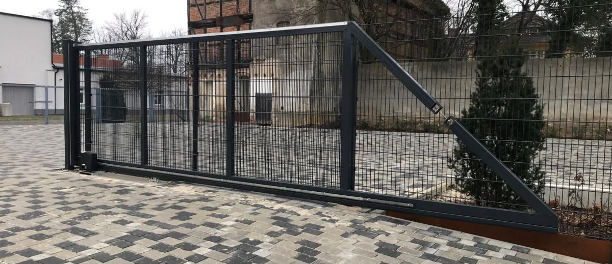 Metalltor und Zaun an einem gepflasterten Eingang, dahinter eine schräge Barriere