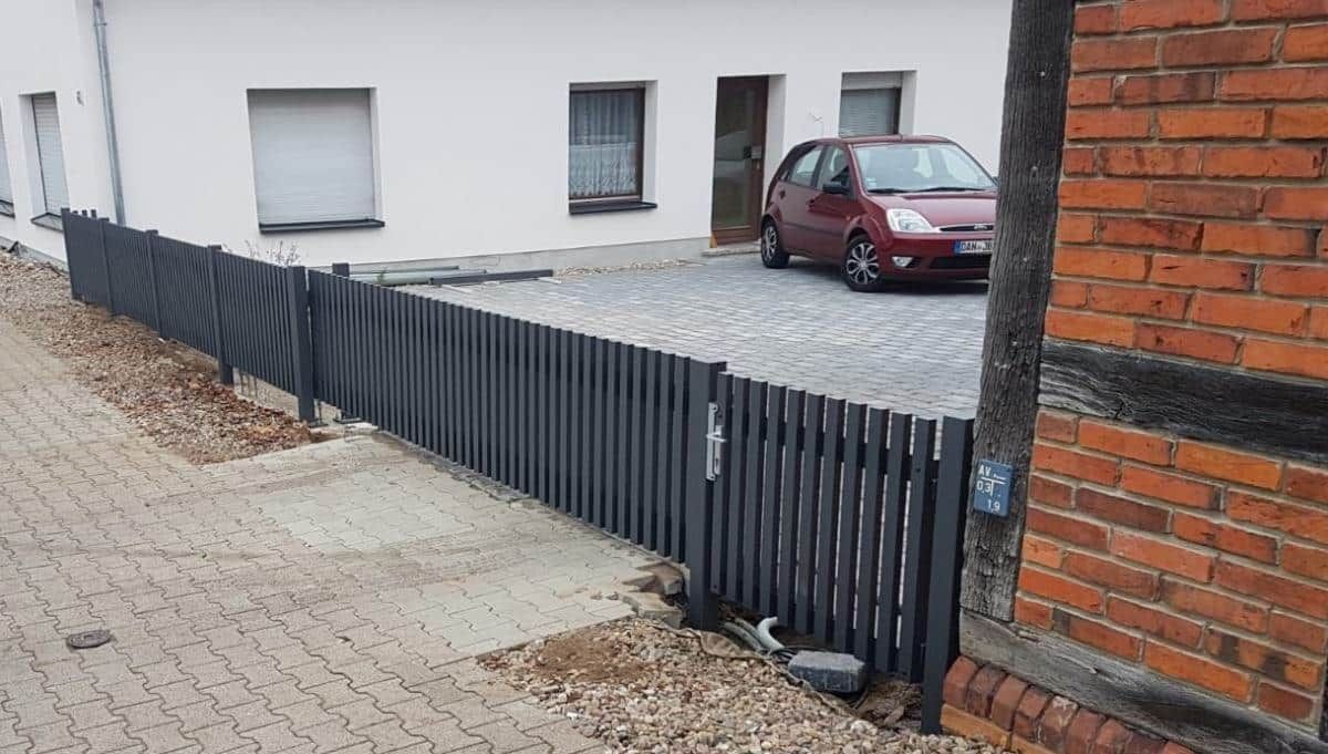 Ein schwarzer Lattenzaun säumt den Gehweg neben einem weißen Gebäude mit einem geparkten roten Auto