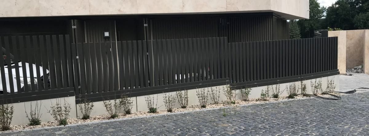 Schwarzer Metallzaun neben einem beigen Gebäude mit gepflasterter Auffahrt und Kiesstreifen