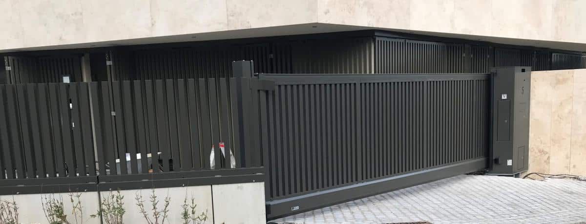 Schwarzes Metallschiebetor mit vertikalen Lamellen entlang einer gepflasterten Auffahrt neben einer beigefarbenen Mauer