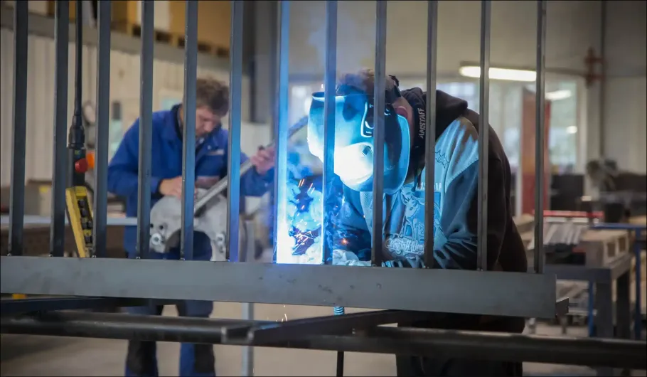 Zwei Arbeiter schweißen in einer Werkstatt einen Metallrahmen zusammen, dabei sprühen hellblaue Funken.