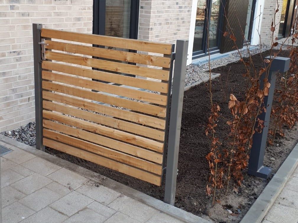 Ein Zaunfeld aus Holzlatten neben einem Backsteingebäude und einem gepflasterten Gehweg
