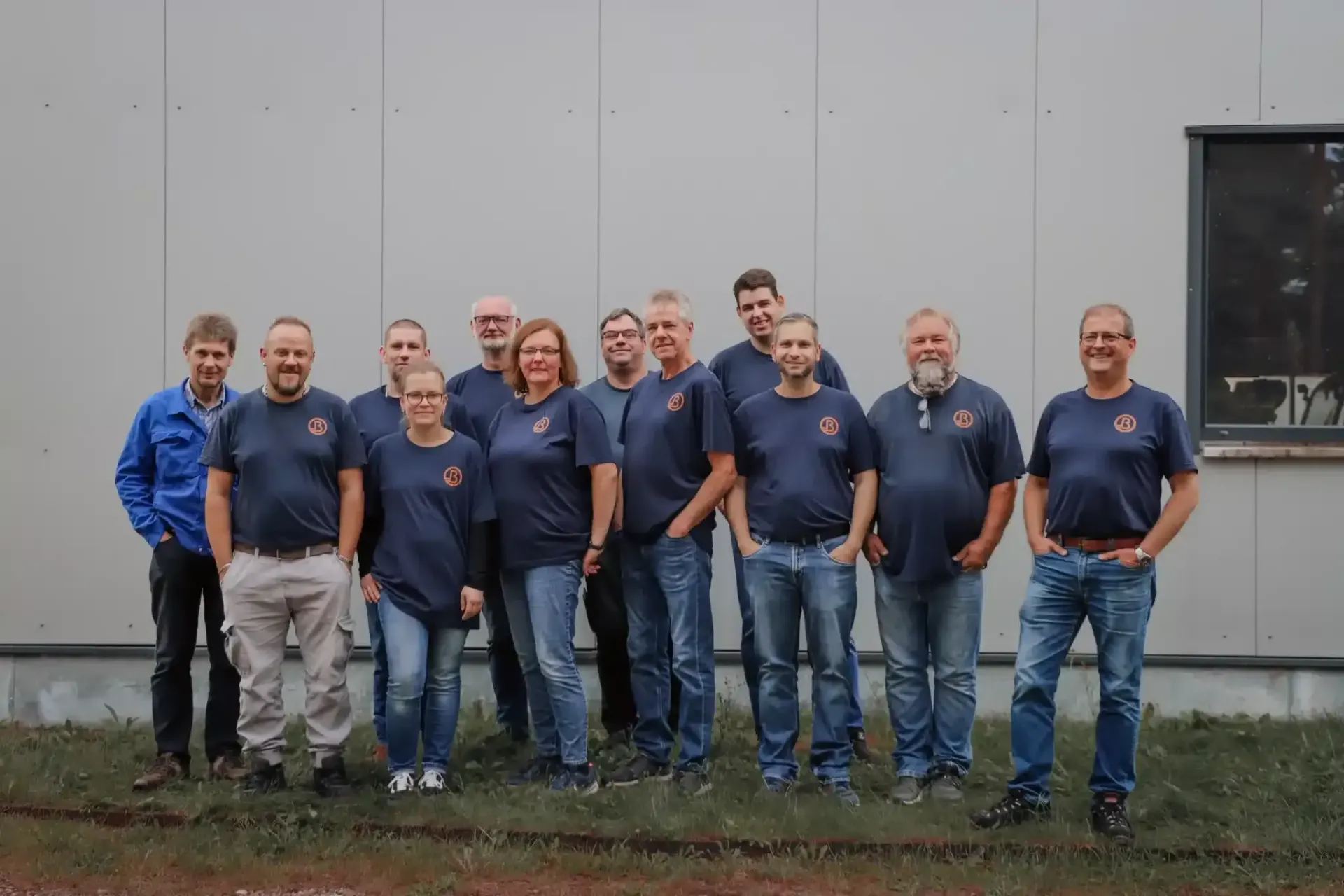 Eine Gruppe von Menschen in blauen Hemden steht im Freien vor einer hellgrauen Wand.