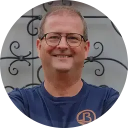 Eine lächelnde Person mit Brille und blauem Hemd posiert vor einem dekorativen Metallzaun.