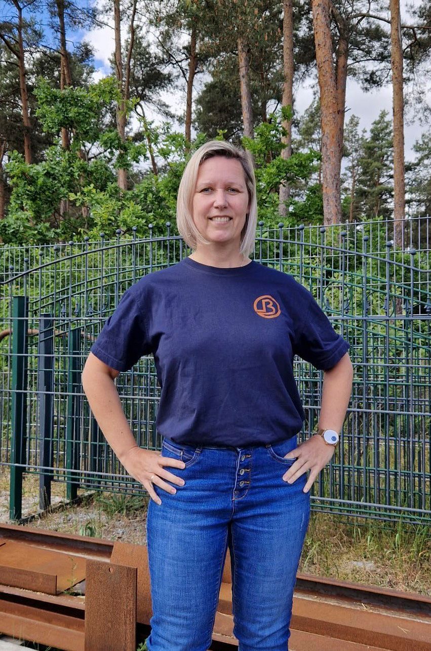 Eine Person steht mit in die Hüften gestemmten Händen im Freien vor einem Zaun und Bäumen