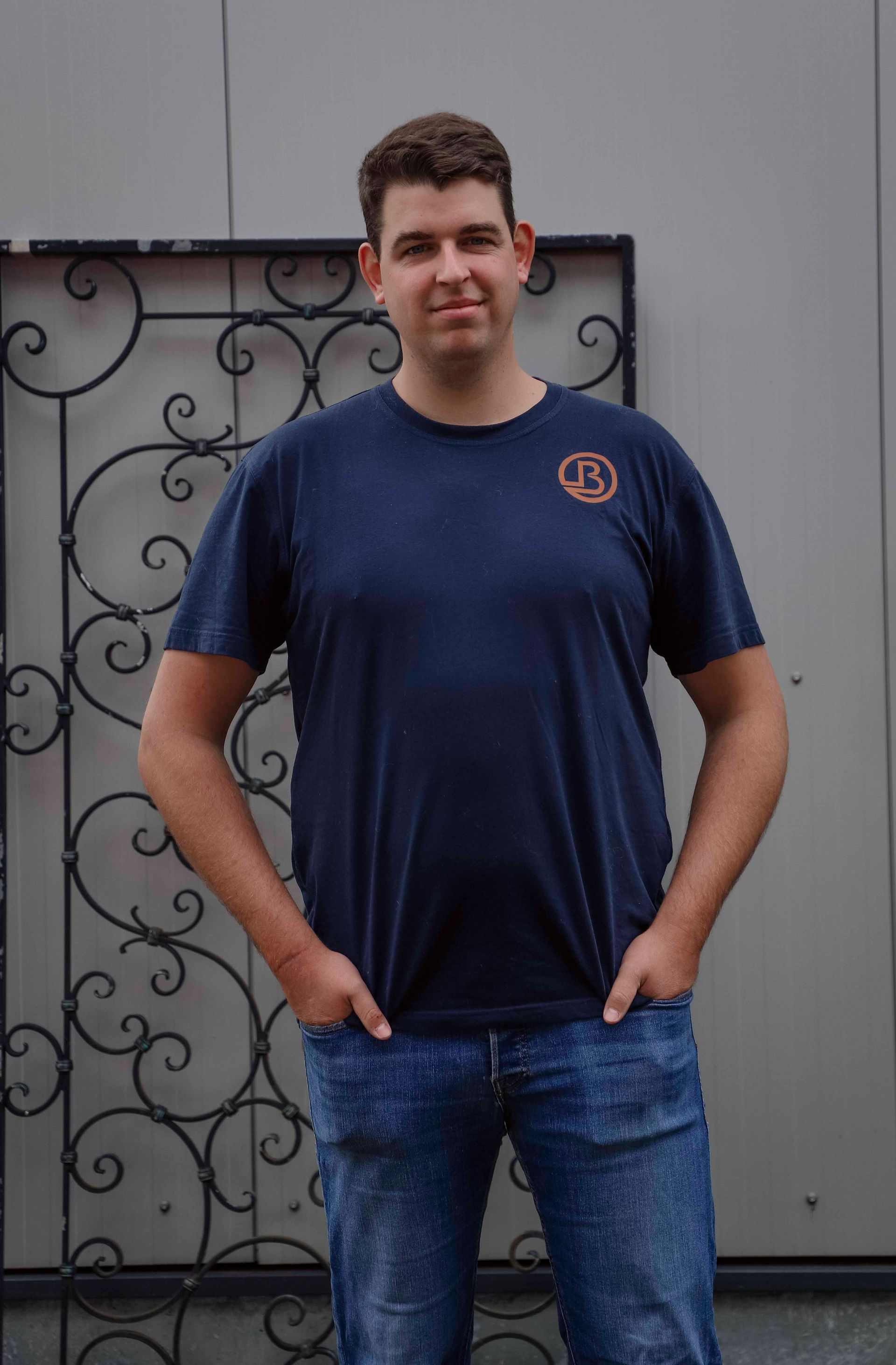 Ein Mann steht im Freien vor einer grauen Wand. Er trägt ein dunkelblaues T-Shirt und Jeans, die Hände in den Taschen.
