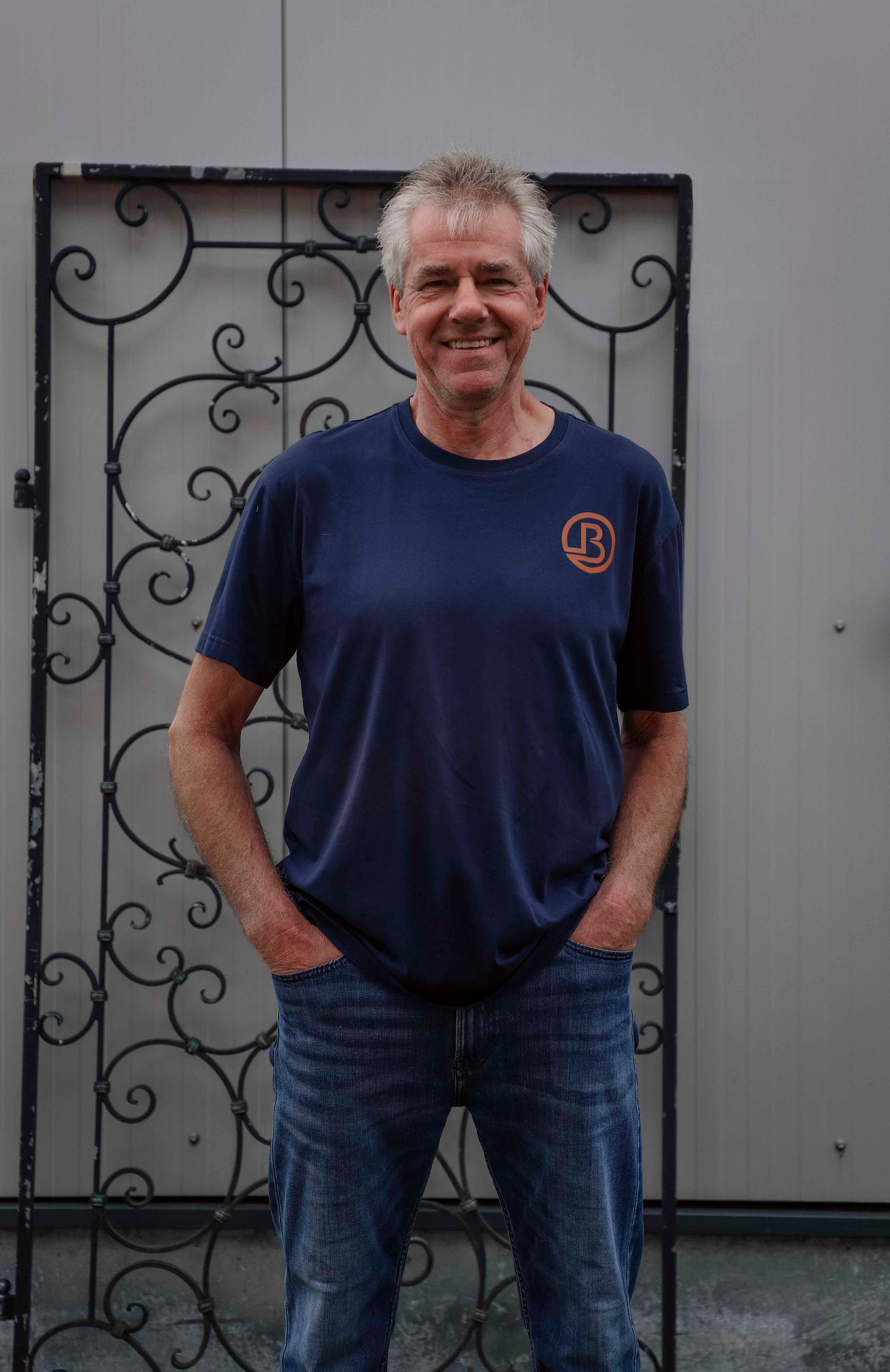 Ein Mann, der im Freien an einem dekorativen Metalltor steht, trägt ein dunkelblaues T-Shirt und Jeans und lächelt.