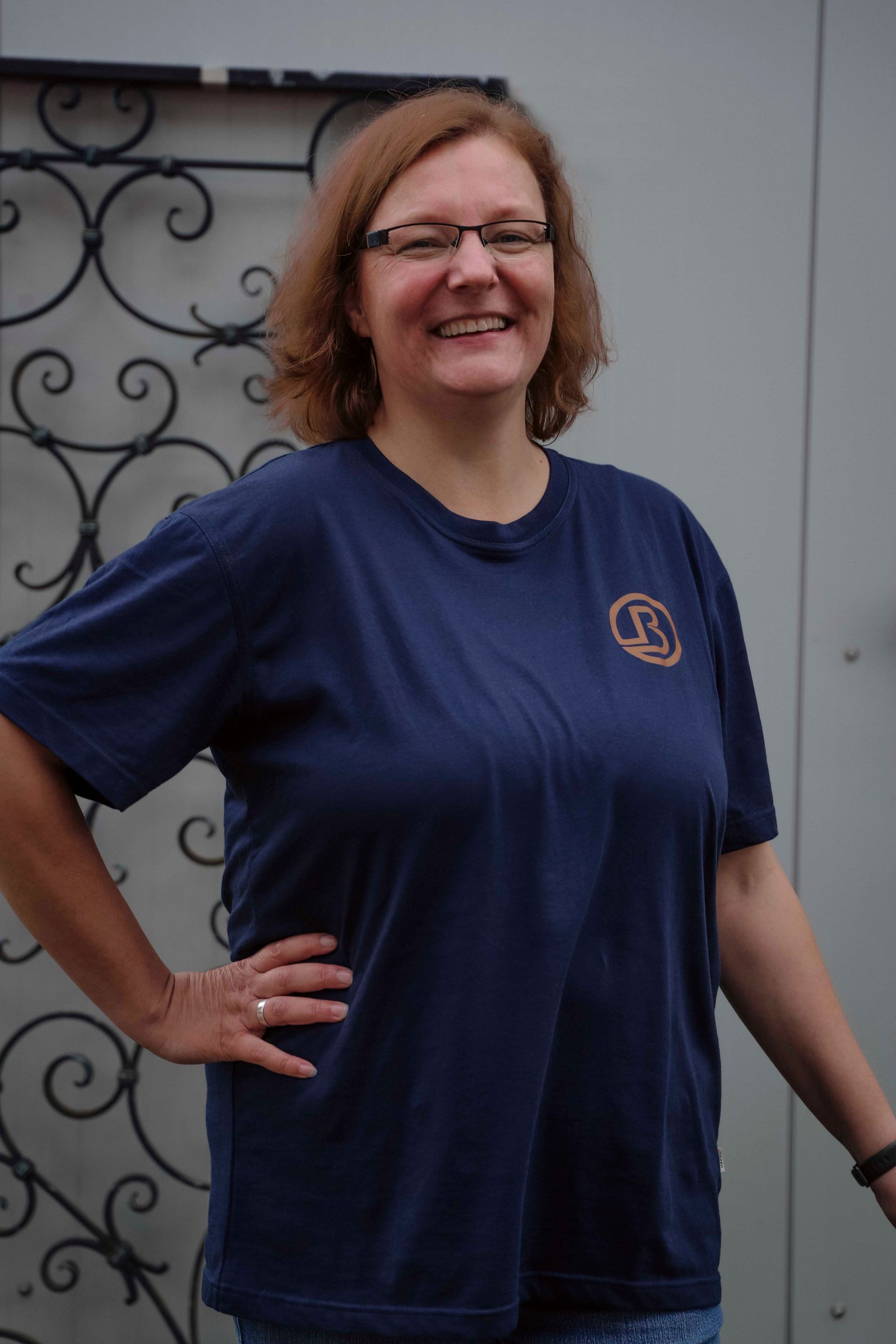 Eine lächelnde Frau in einem dunkelblauen T-Shirt und mit Brille steht mit der Hand in der Hüfte im Freien.