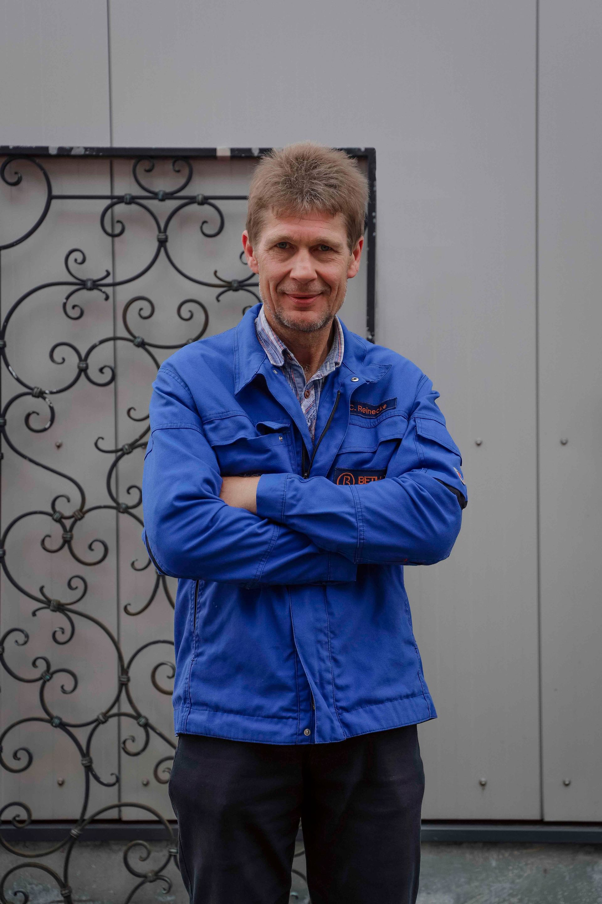 Eine Person in einer blauen Jacke steht mit verschränkten Armen vor einer grauen Mauer und einem schmiedeeisernen Tor.