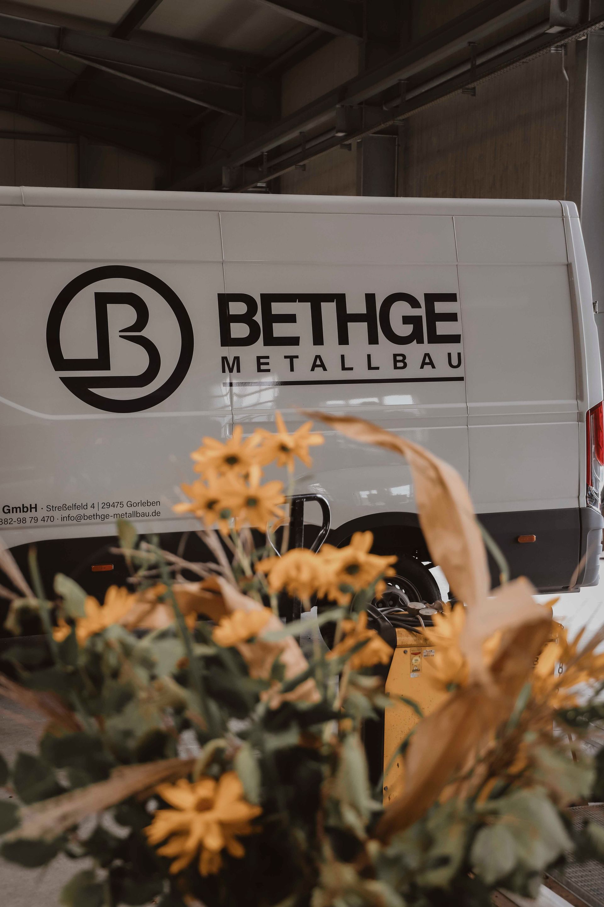 Weißer Anhänger der Firma Bethge Metallbau in einer Lagerhalle, teilweise verdeckt von gelben Blumen im Vordergrund