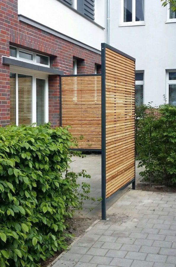 Sichtschutzwand aus Holz neben einem gepflasterten Gehweg, neben einem weißen Backsteingebäude und grünen Sträuchern.