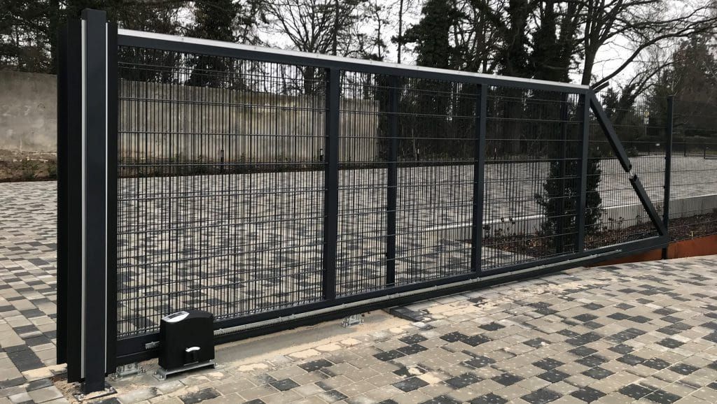 Schwarzes Metallschiebetor entlang einer gepflasterten Auffahrt, im Hintergrund Bäume und ein Zaun.