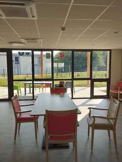 Salle de réunion lumineuse avec tables blanches, chaises rouges et en bois, et grandes fenêtres laissant entrer la lumière du soleil.