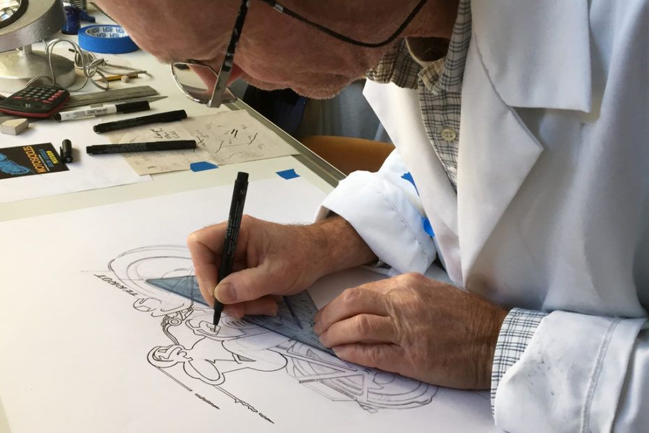 artisan émailleur en blouse blanche dessine avec un stylo noir sur une grande feuille de papier.