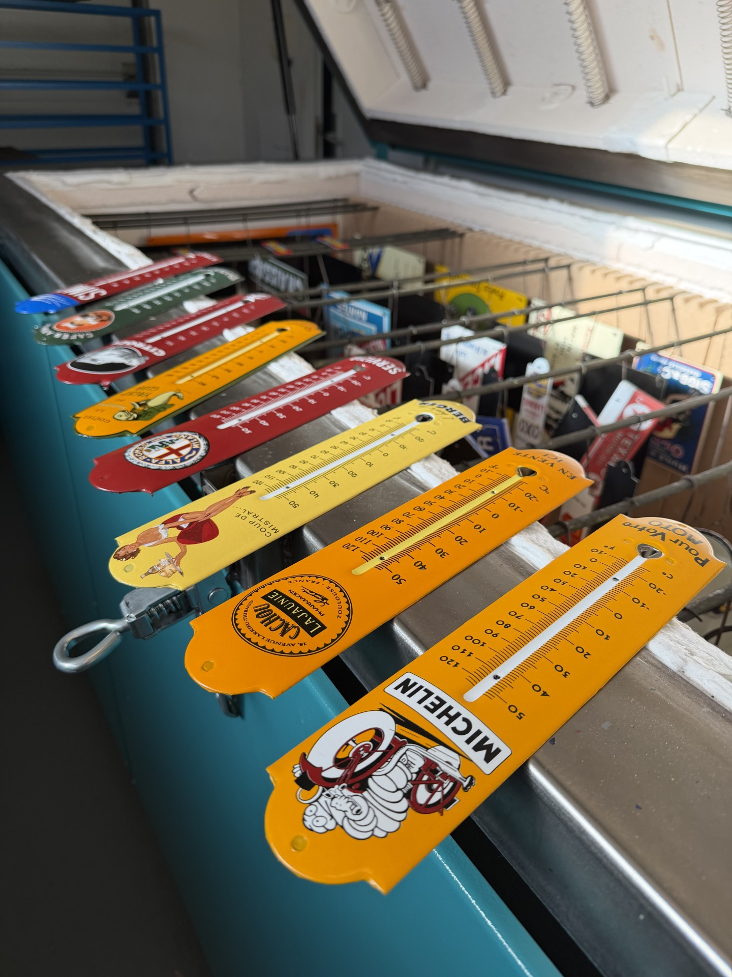 Une rangée de thermomètres vintage colorés, arborant différents logos de marques, est exposée sur un congélateur coffre.