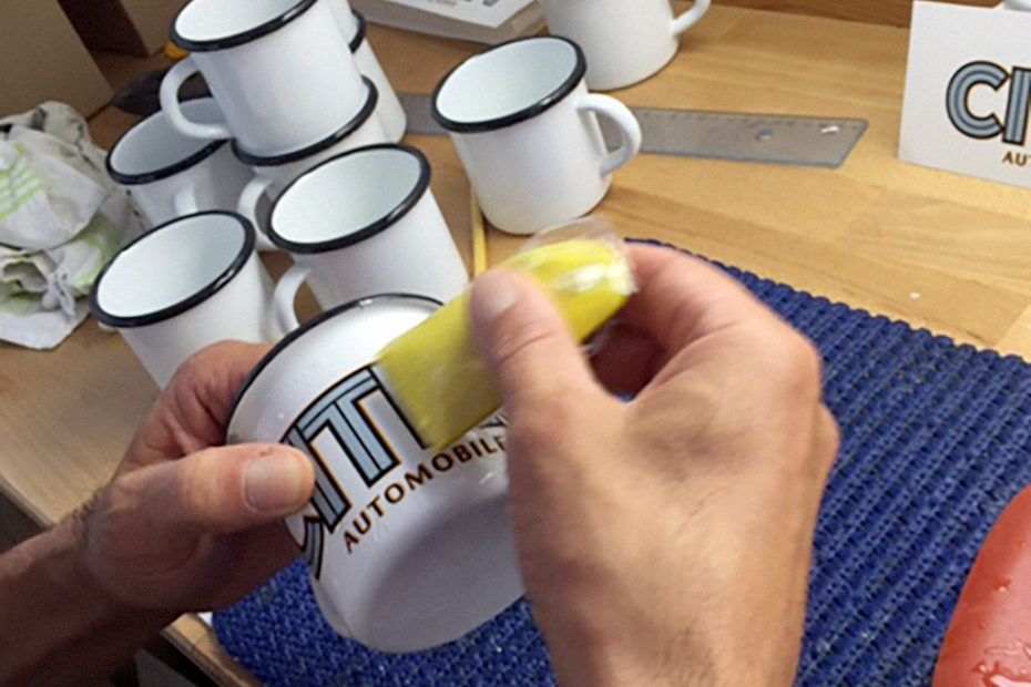 Une personne applique un autocollant sur une tasse en émail blanc, entourée d'autres tasses, sur un tapis bleu.