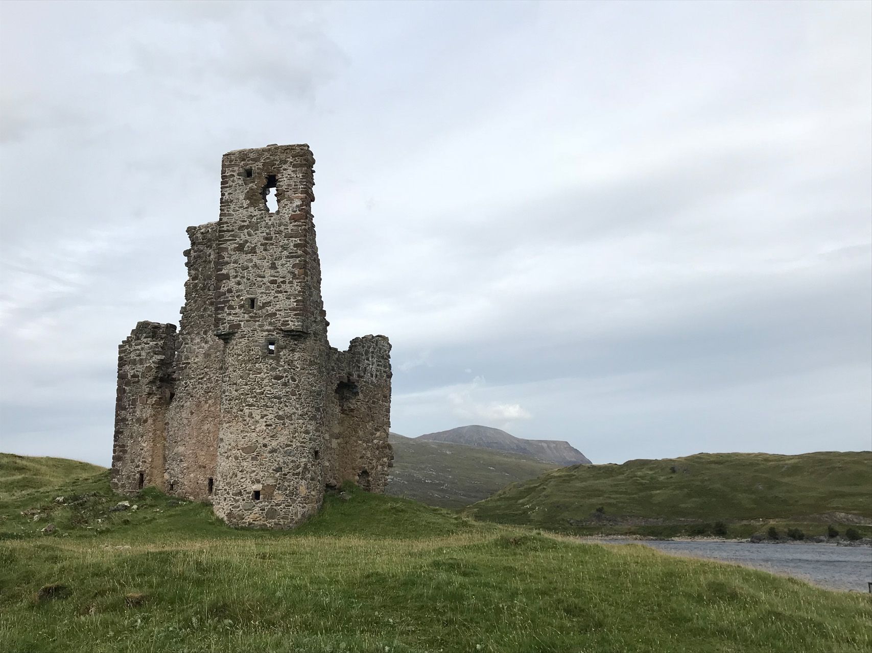 Ein Steinturm steht auf einem grasbewachsenen Hügel neben einem Gewässer.