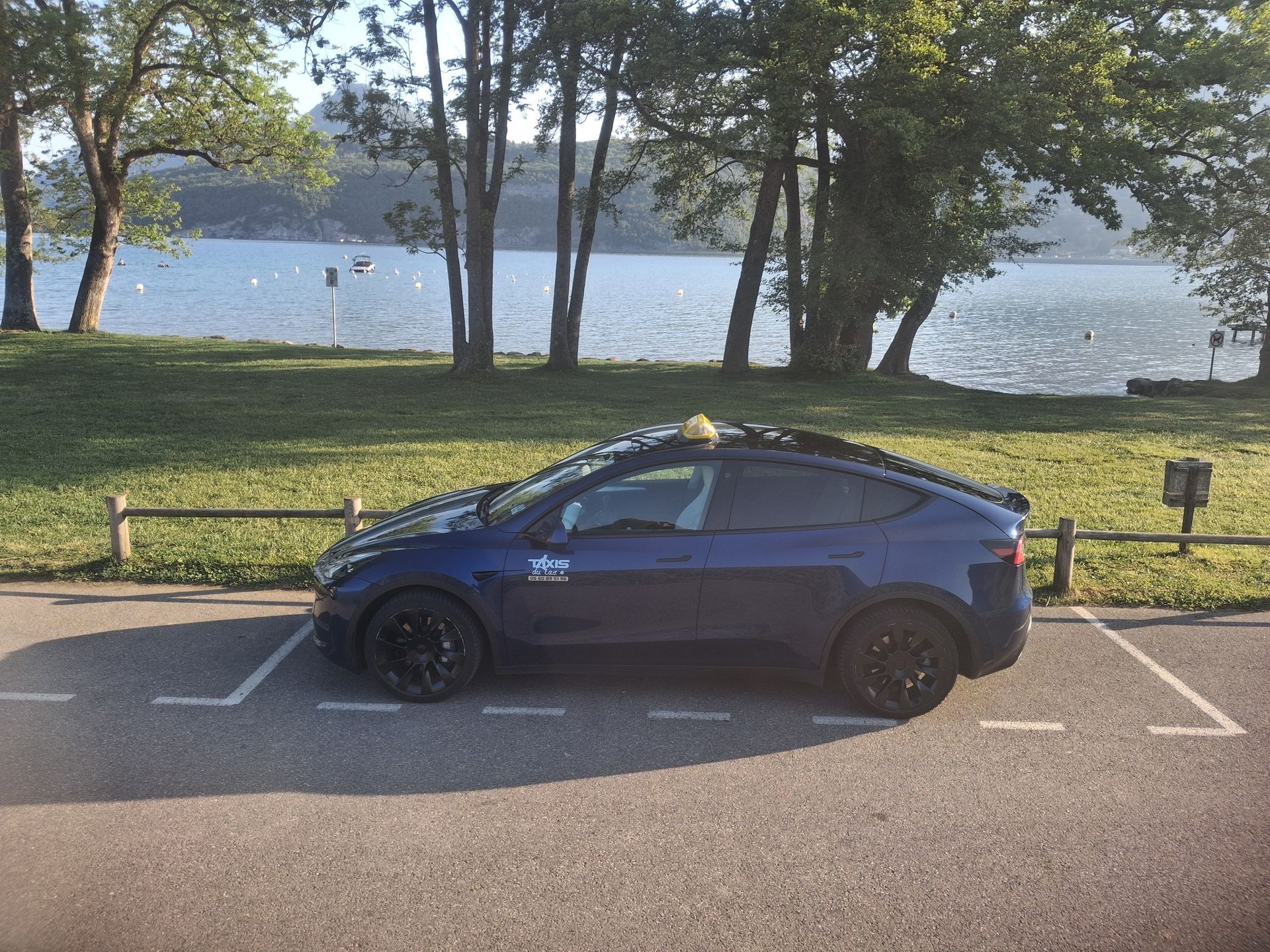 Voiture bleue garée au bord d'un lac, avec des arbres et un banc en arrière-plan.