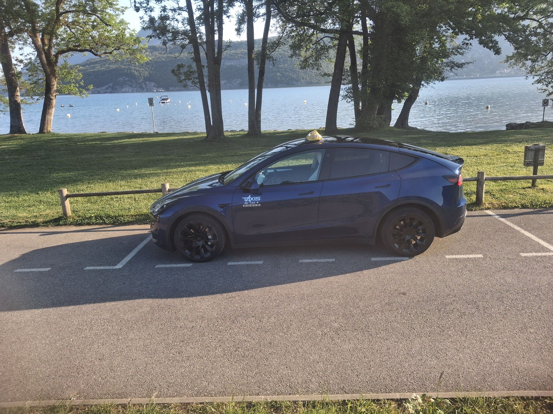 Une voiture à hayon bleu foncé garée au bord d'un lac, avec des arbres et du soleil en arrière-plan.