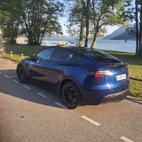 Une berline Tesla bleue est garée au bord d'une route longeant un lac, avec des arbres et des montagnes en arrière-plan.