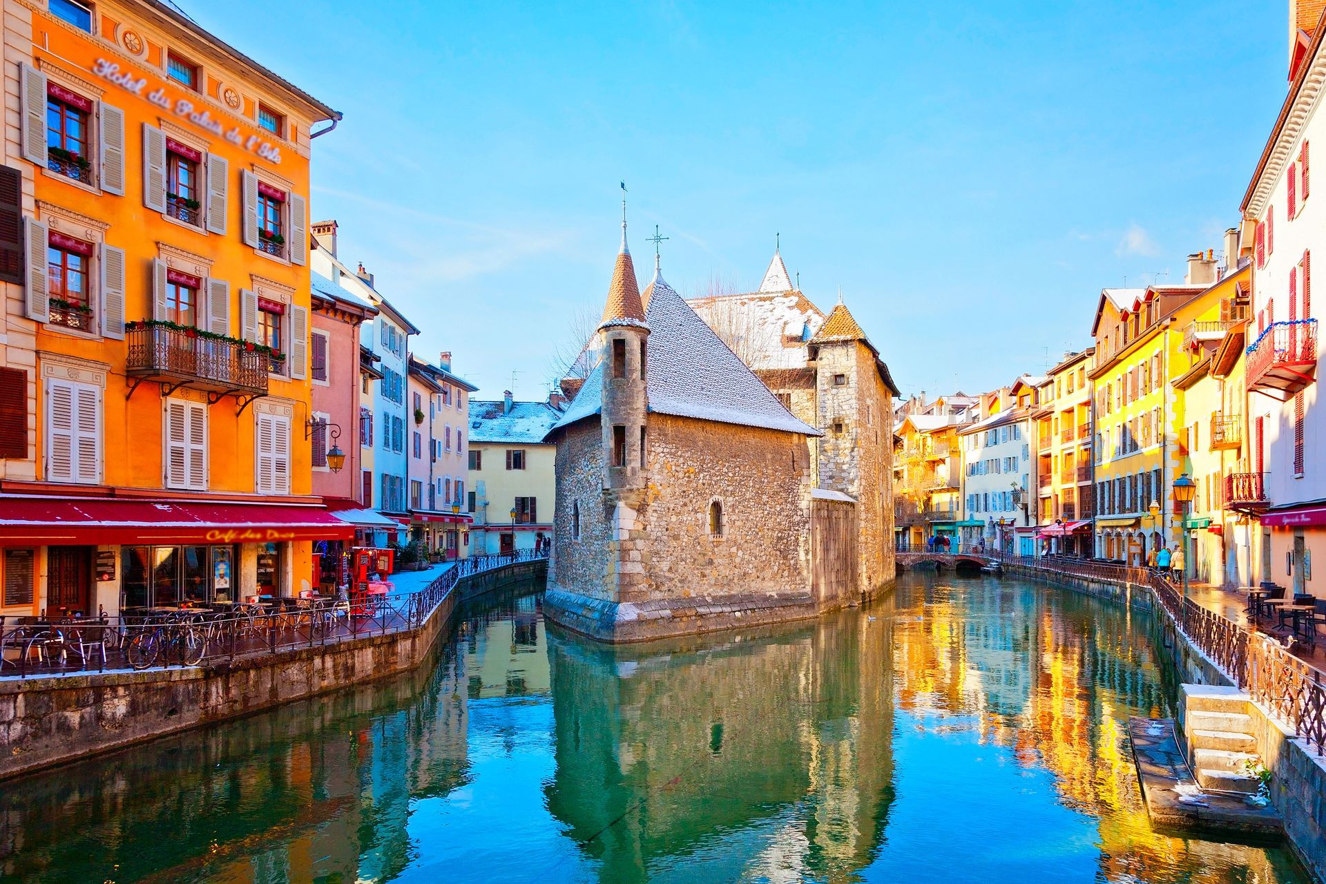 Des bâtiments colorés bordant le canal et une tour en pierre se reflétant dans l'eau calme sous un ciel bleu.