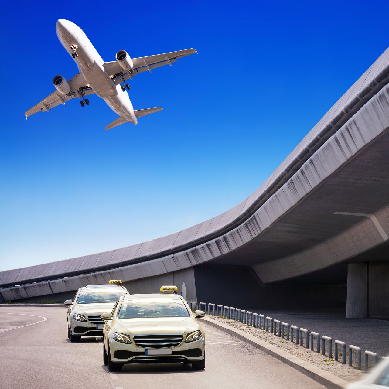 Deux voitures roulent le long d'un mur en béton tandis qu'un avion de ligne survole la zone, se détachant sur un ciel bleu.