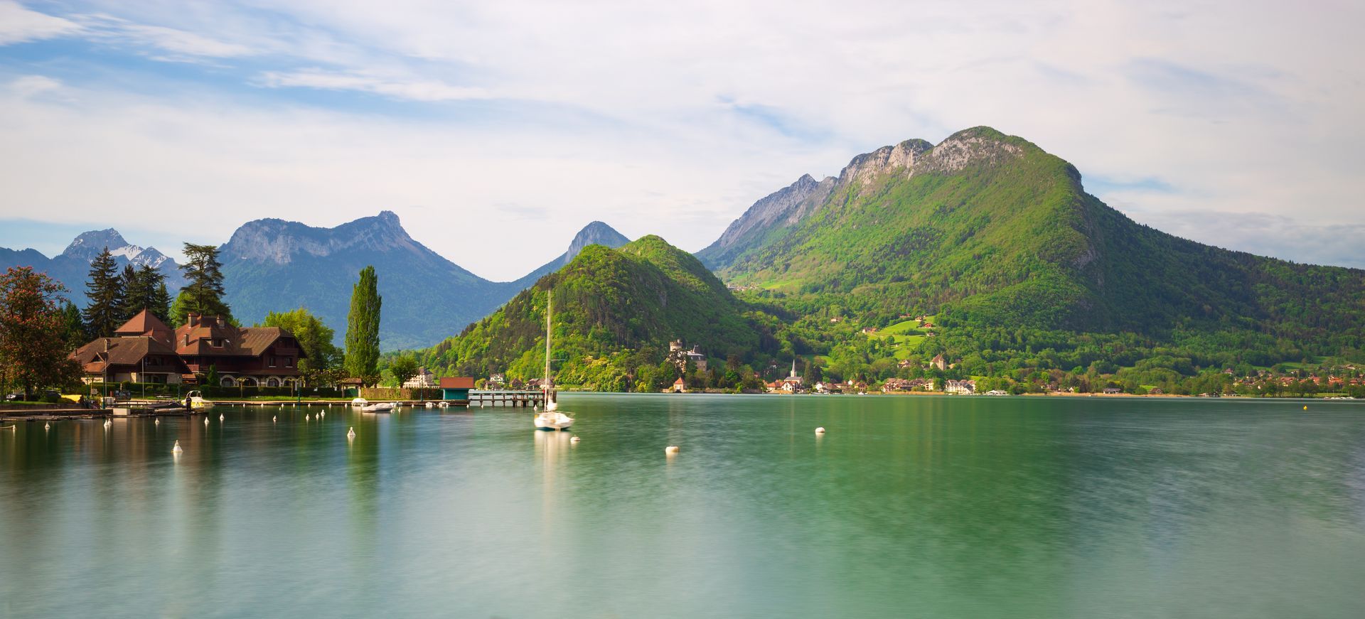 Lac serein avec un petit voilier, des montagnes et des collines verdoyantes sous un ciel lumineux.