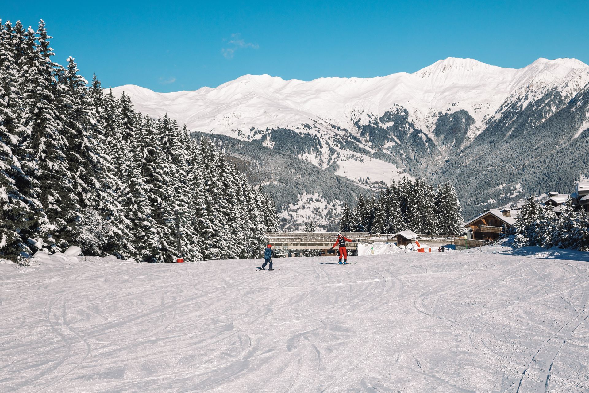 Piste de ski enneigée avec quelques skieurs, des pins et des sommets montagneux sous un ciel bleu.
