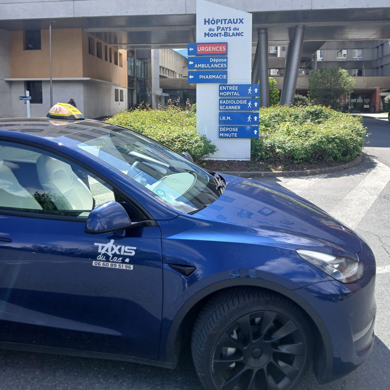 Un taxi bleu garé devant l'entrée d'un hôpital, à côté d'un panneau et d'arbustes.