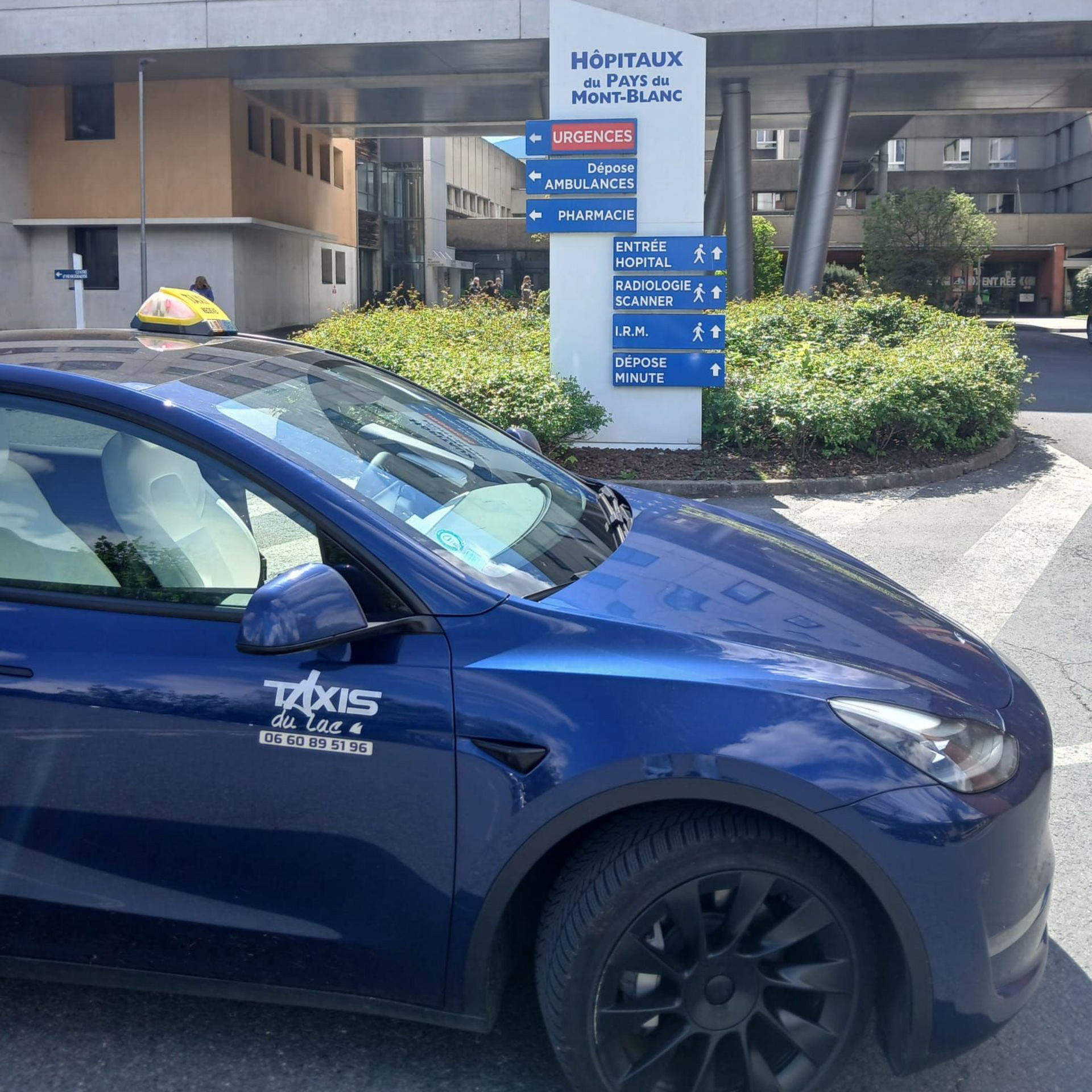 Un taxi bleu est stationné devant l'entrée d'un hôpital, avec un panneau et des buissons devant.