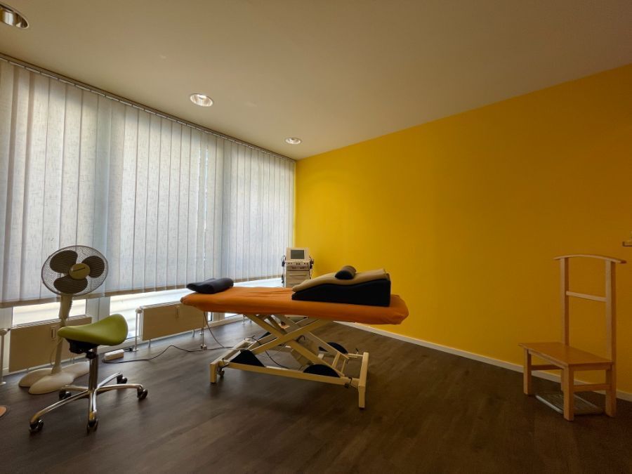 Therapieraum mit Behandlungstisch, Ventilator und gelber Akzentwand.