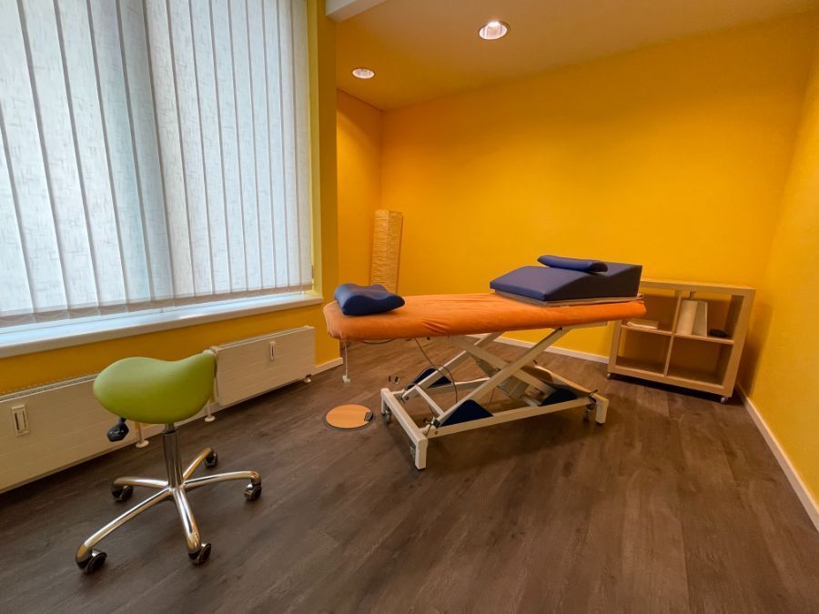 Therapieraum mit orangefarbenen Wänden, Massagetisch, grünem Stuhl und Fenster mit Jalousien.