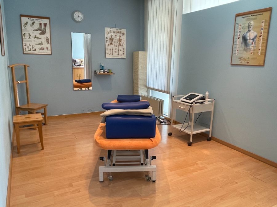 Therapieraum mit verstellbarem Massagetisch, medizinischer Ausrüstung, anatomischen Diagrammen und blauen Wänden.