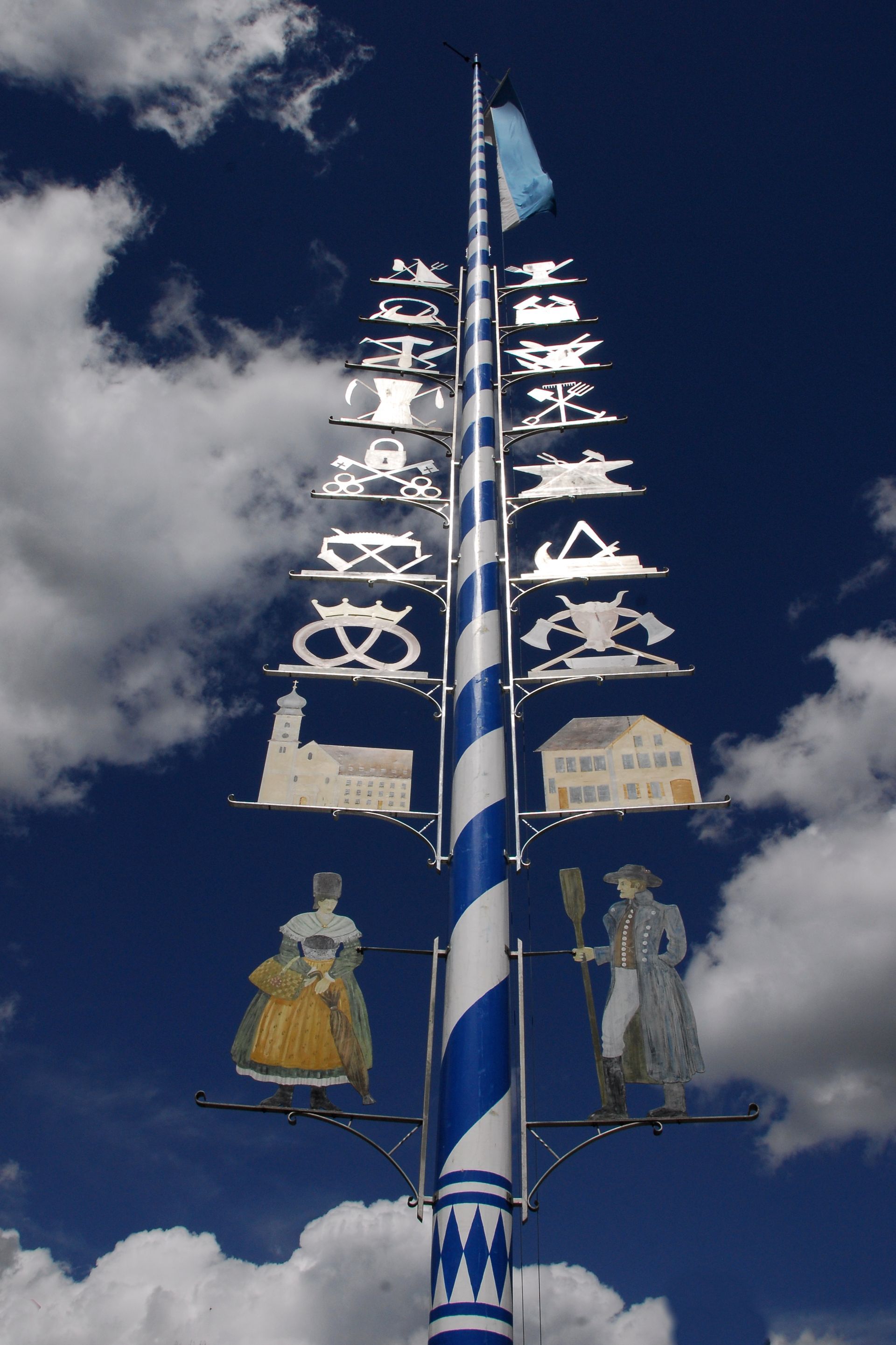 Ein blau-weiß gestreifter Fahnenmast mit einer Frau und einem Mann, sowie weiteren Symbolen daran befestigt
