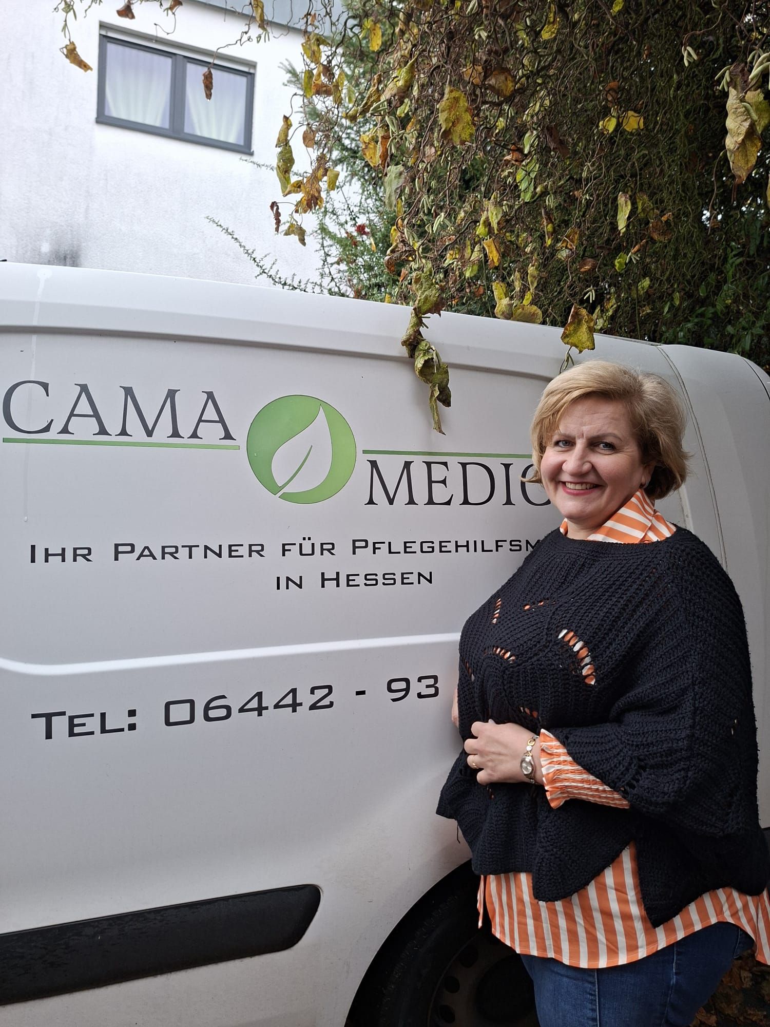 Frau steht neben einem weißen Lieferwagen mit dem Logo „CAMA MEDIC“; Telefonnummer sichtbar. Herbstlaub im Hintergrund.