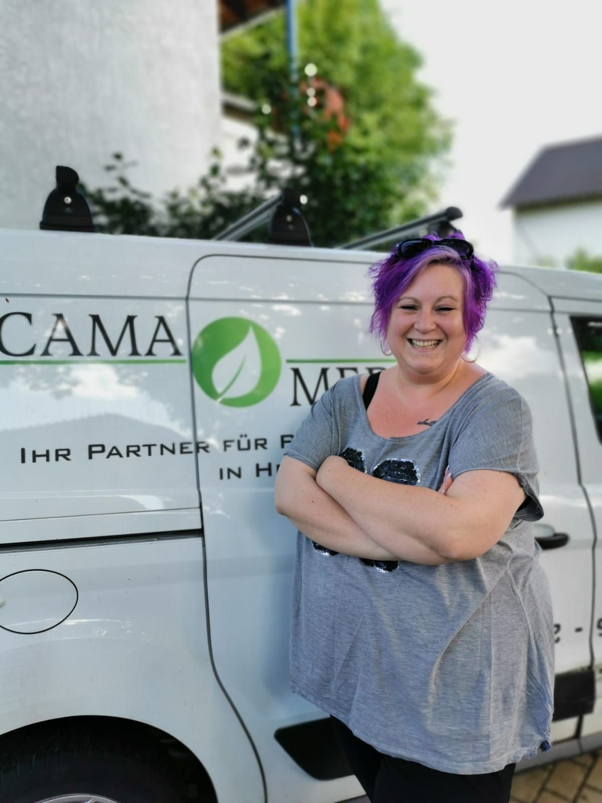 Frau mit lila Haaren lächelt mit verschränkten Armen neben einem weißen Lieferwagen mit grünem Logo „CAMA MED“.