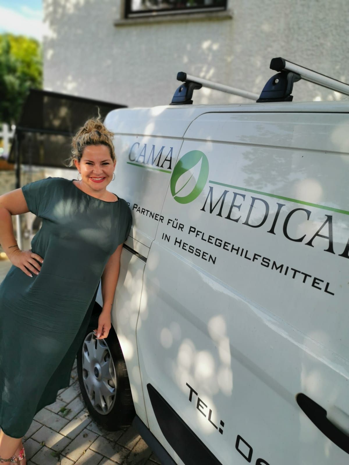 Eine Frau in einem grünen Kleid posiert neben einem weißen Lieferwagen mit dem Logo „CAMAN MEDICAL“.