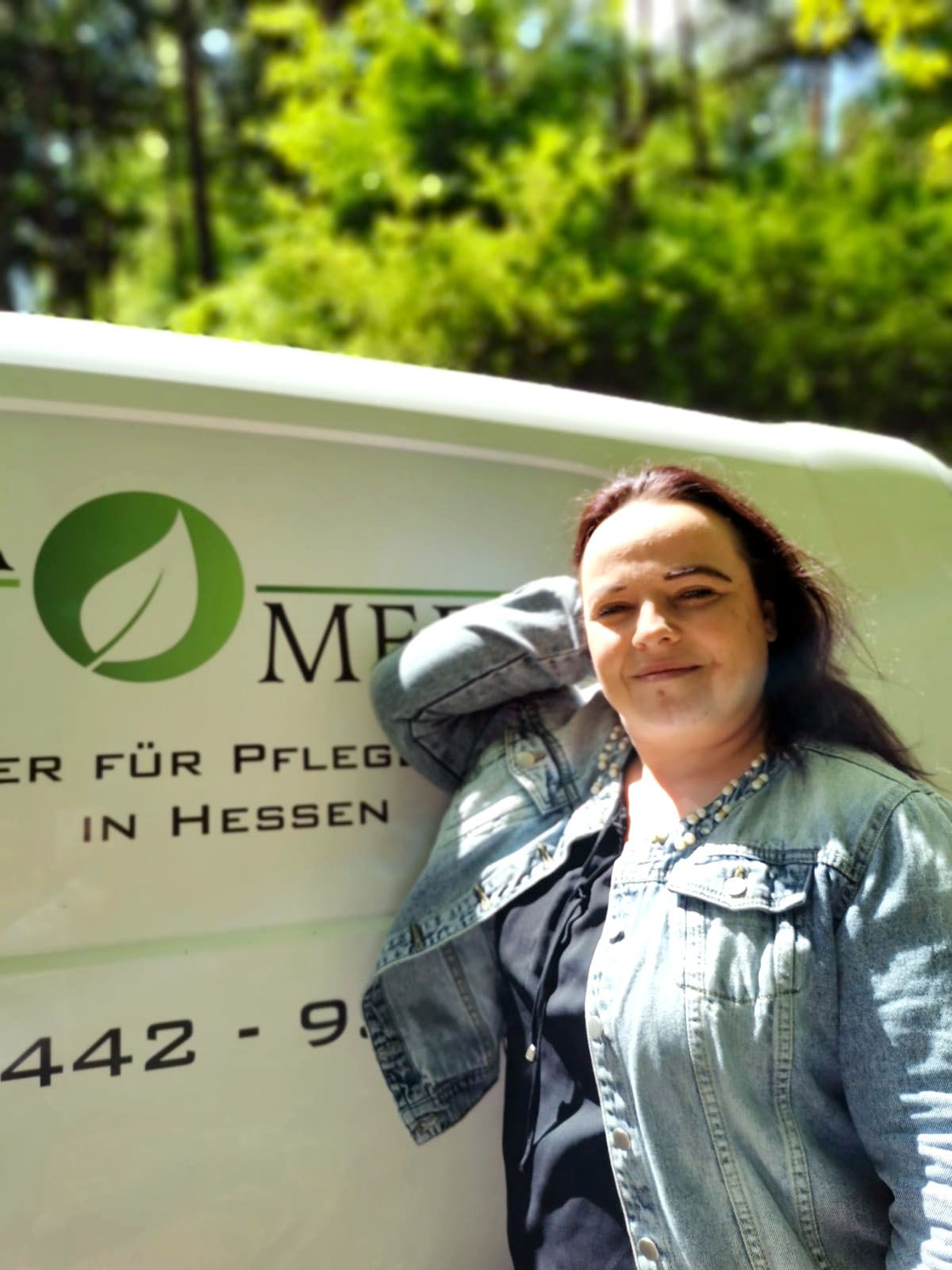 Eine Frau in einer Jeansjacke lehnt im Freien an einem weißen Lieferwagen mit grünem Logo.