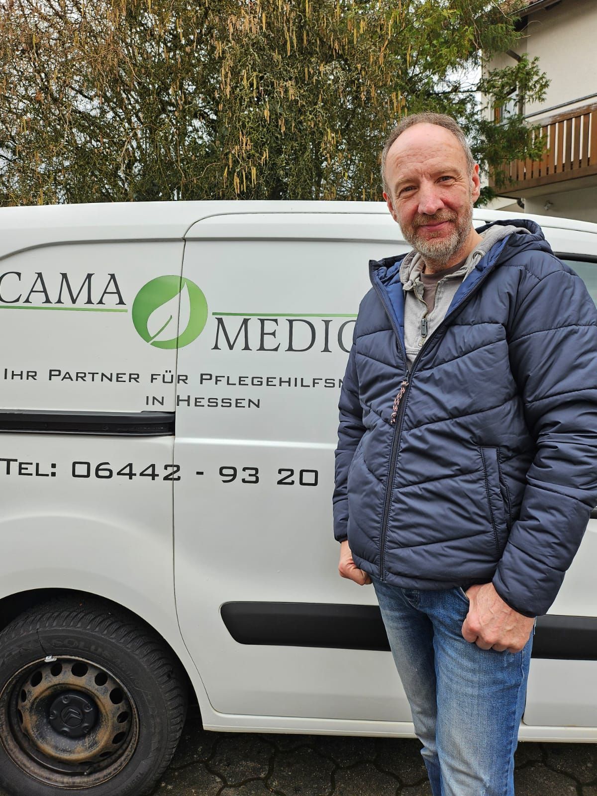 Ein Mann steht neben einem weißen Lieferwagen mit dem Logo „CAMA MEDIC“. Er trägt eine blaue Jacke und Jeans.