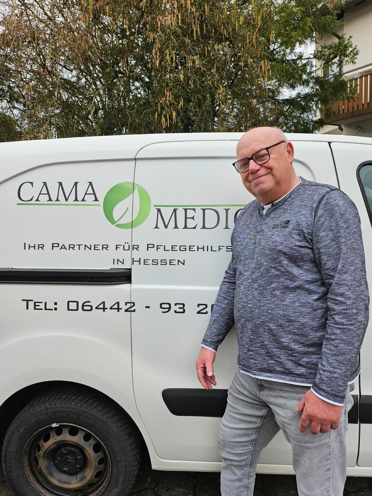 Ein Mann steht neben einem weißen Lieferwagen mit dem Logo. Er lächelt, trägt eine Brille und einen blauen Pullover.
