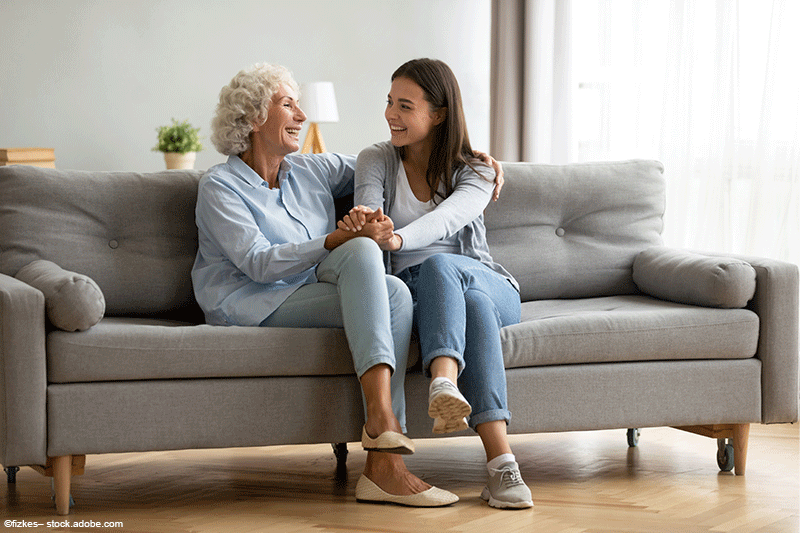Eine Frau und ein älterer Erwachsener sitzen Händchen haltend und lächelnd auf einem grauen Sofa im Wohnzimmer.