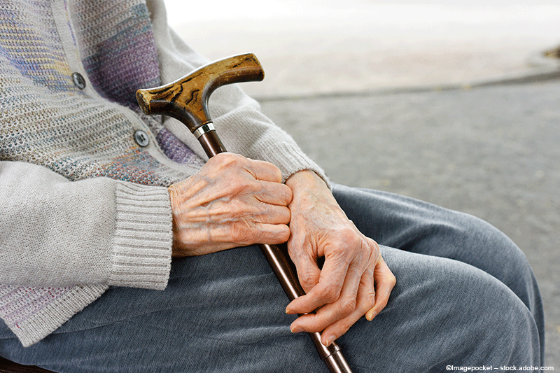 Die Hände einer Person halten einen Gehstock, der auf dem Schoß ruht; sie trägt einen hellen Pullover.