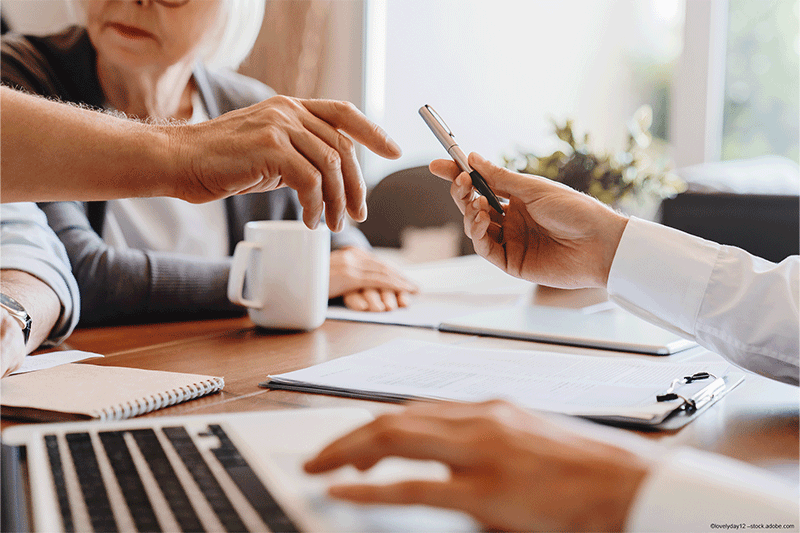 Hände streichen mit einem Stift über Dokumente auf einem Tisch; eine andere Person tippt auf einem Laptop.