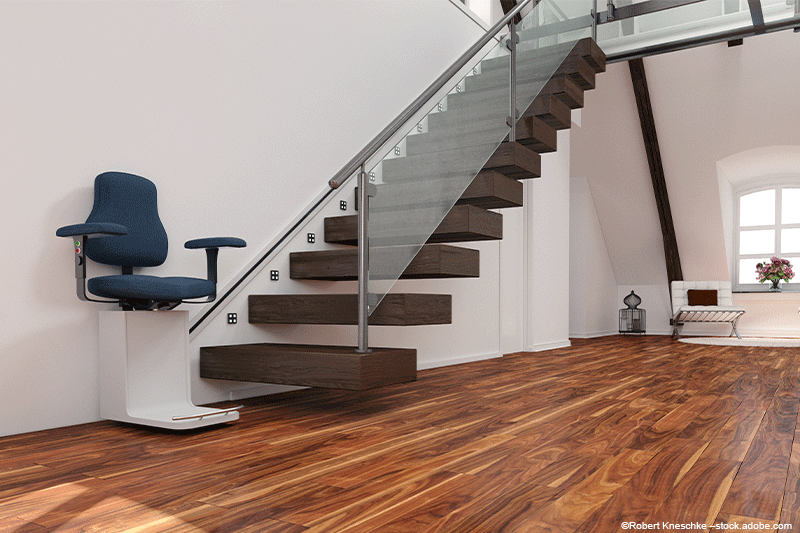 Treppenlift auf einer freitragenden Treppe mit Glasgeländer. Blauer Sitz, Parkettboden, weiße Wände.