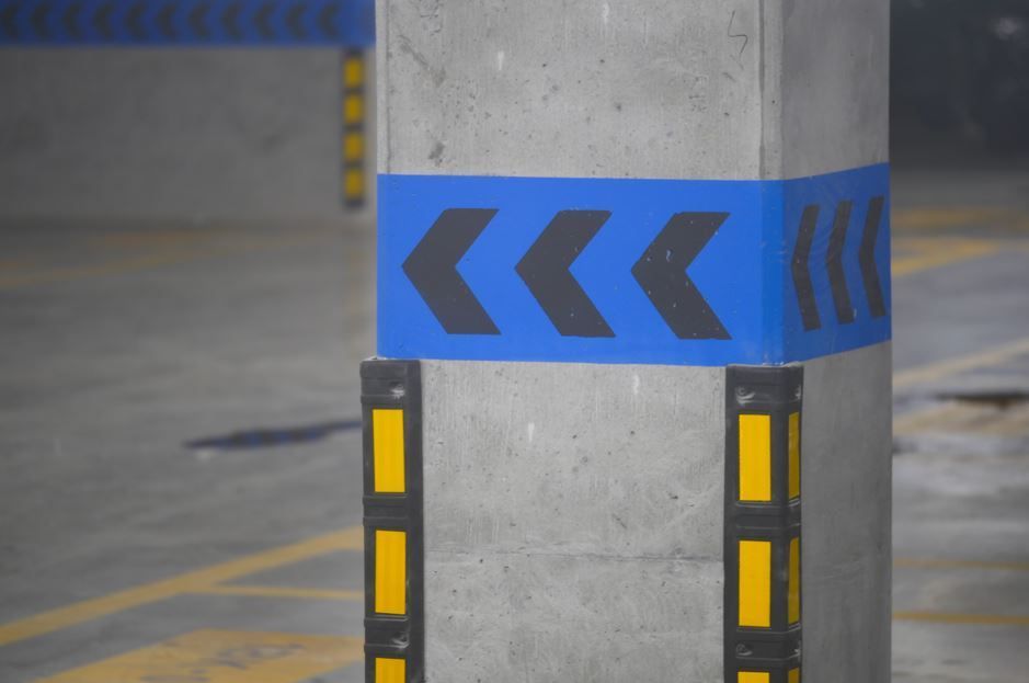Pilar de hormigón en estacionamiento con cinta de chevron azul y negra y protectores de esquinas amarillos y negros.
